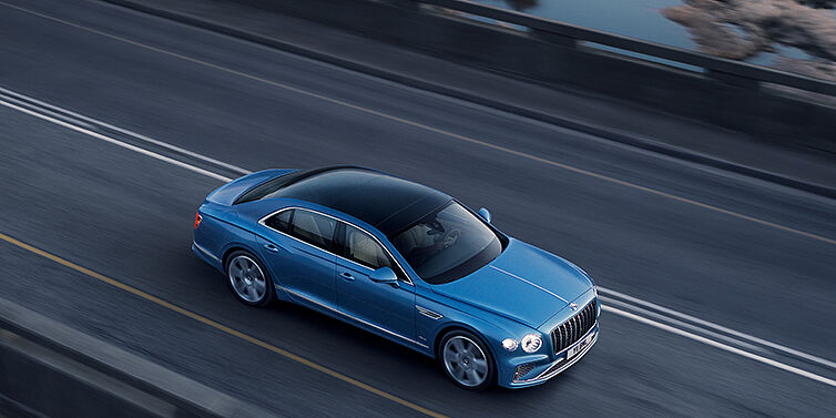 Bentley Budapest New Bentley Flying Spur Azure sedan in Light windsor blue paint by Mulliner, with 22 inch Azure wheels seen from overhead driving dynamically on a country road